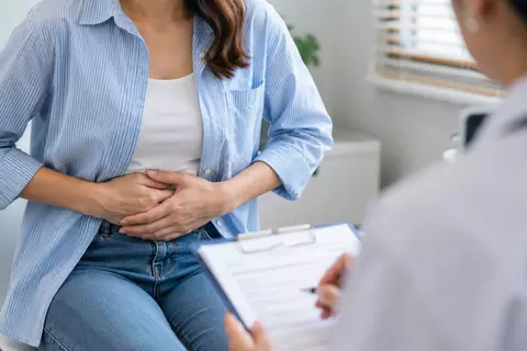 Frau hält sich beim Arztbesuch den Bauch und spricht über Blähungen und Bauchschmerzen.