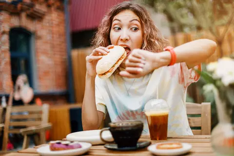 Junge Frau isst hastig im Restaurant und schaut auf die Uhr – schnelles Essen kann Blähungen begünstigen.