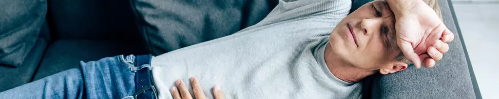 mann-liegt-mit-hand-auf-dem-kopf-auf-dem-sofa-hält-sich-hände-auf-den-bauch-gastritis-bauchschmerzen-voellegefuehl-magenbrennen-gaspan